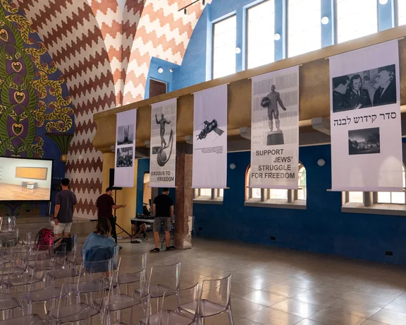 Intérieur d’une salle d’exposition avec des banderoles détaillant l’art juif et les luttes pour la liberté, comprenant un écran vidéo et des rangées de chaises transparentes vides.