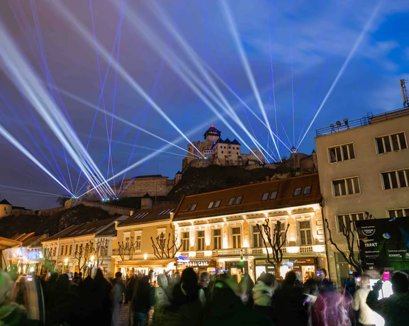 Vue nocturne spectaculaire du château de Trenčín illuminé par des faisceaux lasers blancs et bleus lors d'un spectacle public, avec une foule observant en contrebas.
