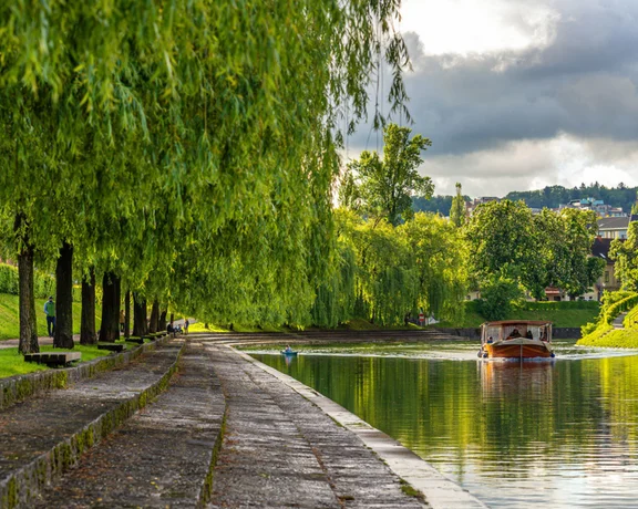 Tree-lined riverbank with a small boat on calm water under a cloudy sky.