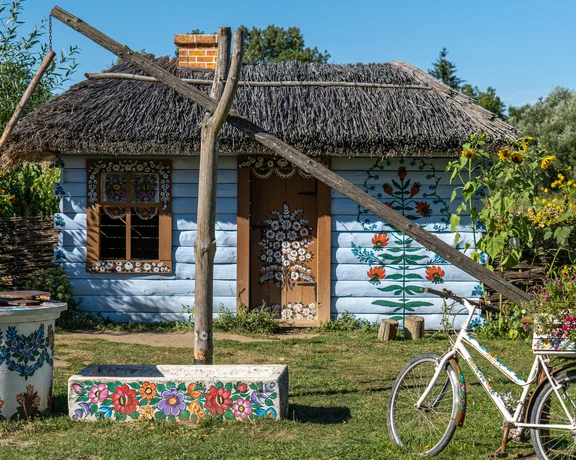 Thatched cottage with painted flowers, an old well, and a white bicycle in a sunny garden.