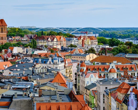 Panoramic view of a historic European city with red-tiled roofs, a brick Gothic church, and bridges over a river