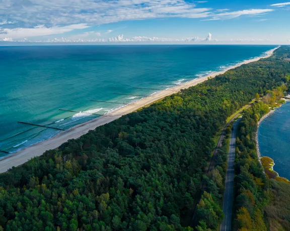 Aerial view of a narrow forested peninsula between blue sea and lagoon under a partly cloudy sky.