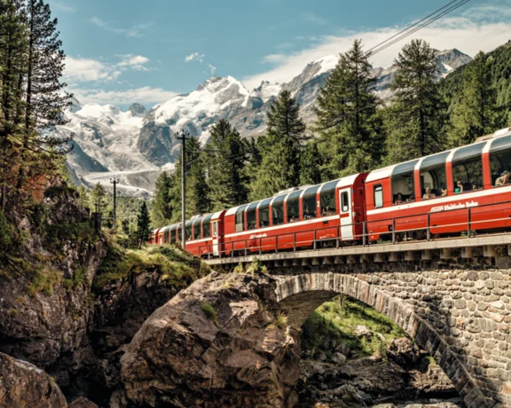 Red train crosses a stone bridge in a mountainous forest with snowy peaks in the background.