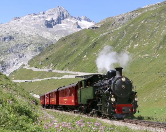 Steam train traveling through green mountains with snow-capped peaks in the background.