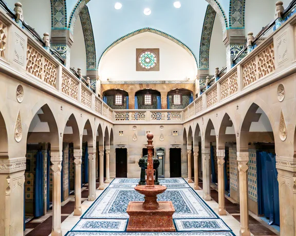 Interior of a traditional bathhouse with arches, tiled walls, and a central fountain.