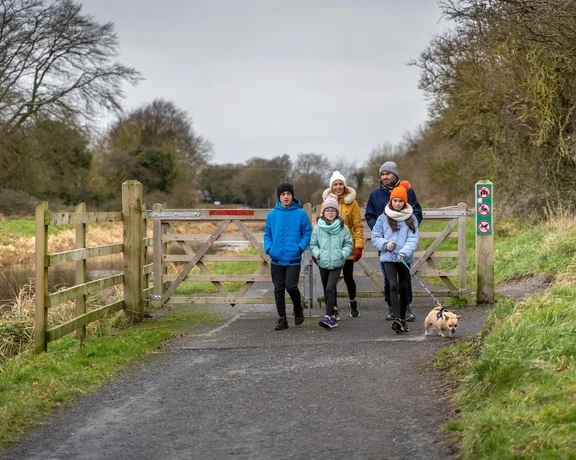 Family and small dog walking on a country path near a wooden gate on a cloudy day.