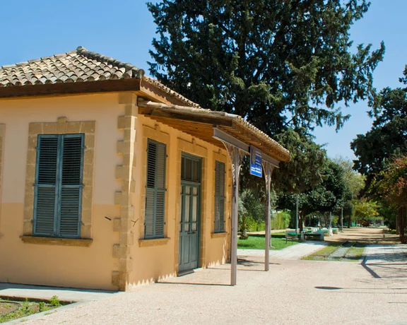 Small yellow building with green windows beside a path, surrounded by trees under a blue sky.