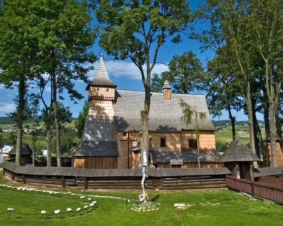 Old wooden church surrounded by green trees and grassy landscape under a blue sky.