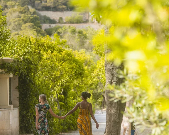 Two women in colorful dresses holding hands and walking down a sunny, leafy path.