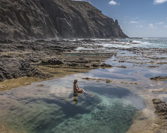 Person relaxing in a tide pool by rocky cliffs and the ocean under a blue sky.