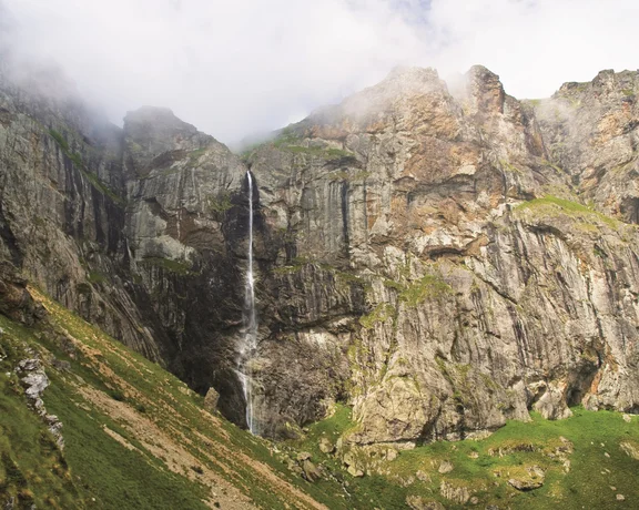 Tall waterfall cascading down a rocky mountain cliff with clouds and green grass.
