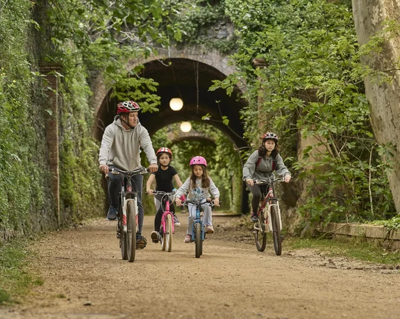 Family of four riding bikes on a forest path under a stone tunnel.
