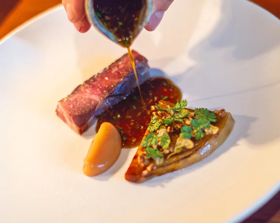 Sauce being poured over seared steak, vegetables, and puree on a white plate.