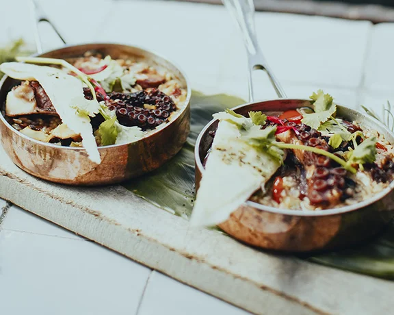 Two bowls of grilled octopus with vegetables and flatbread on a wooden tray.
