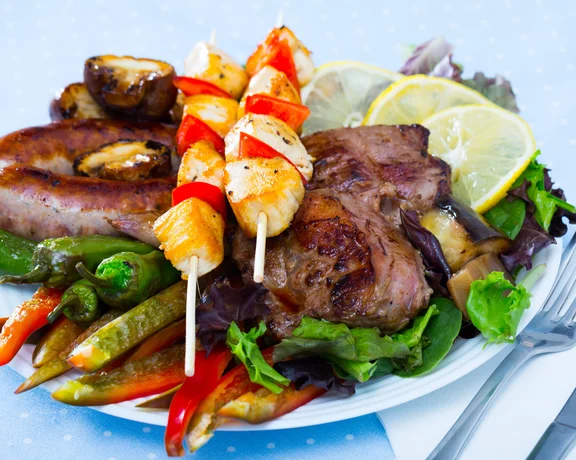 Grilled meat, sausage, and vegetable skewers served with mixed vegetables and lemon slices.