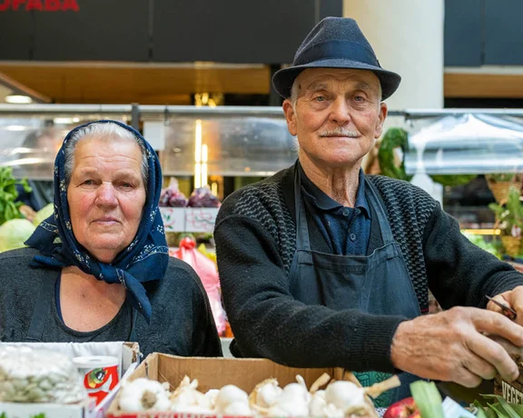 Elderly couple selling vegetables at an indoor market stall, smiling warmly.