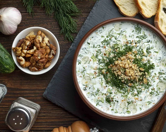 Bowl of creamy dip with herbs and walnuts, surrounded by bread, cucumber, and garlic.