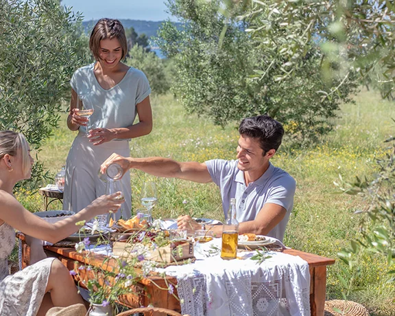 Three people enjoying an outdoor meal at a table in a sunny, green garden.
