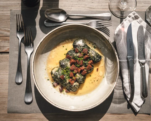 A bowl of spinach gnocchi with sauce and diced toppings on a wooden table setting.