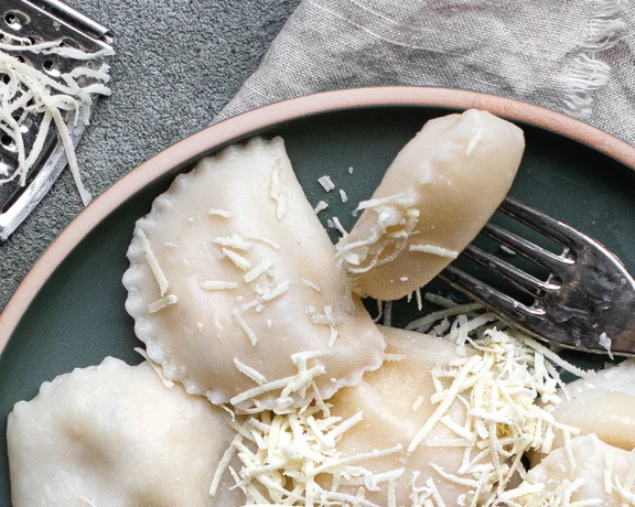 Ravioli topped with grated cheese on a dark plate, fork, and a block of cheese nearby.