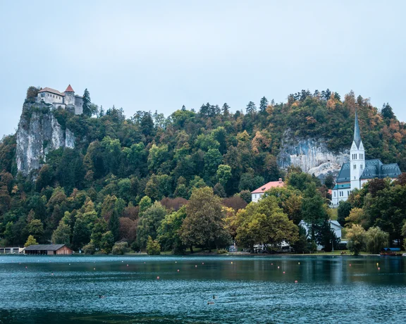 Castle on a forested hill above a lake with a church and autumn trees.