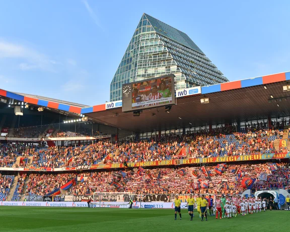 Soccer teams enter a crowded stadium with a glass pyramid building in the background.