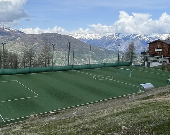 Small green soccer field on a hillside with mountains and buildings in the background.