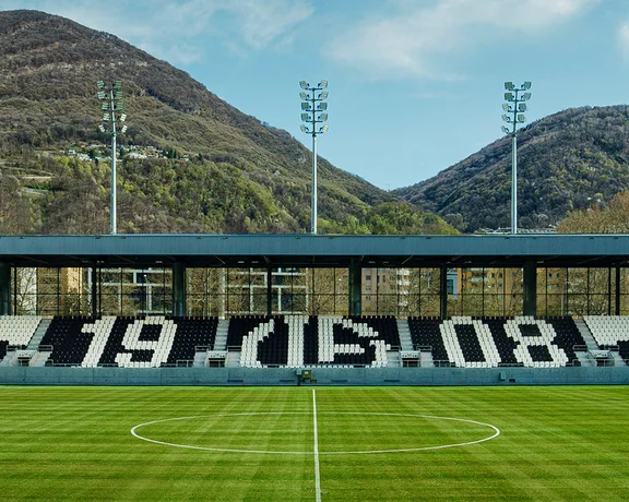 Soccer stadium with large numbers "1908" on seats, green field, mountains in background.