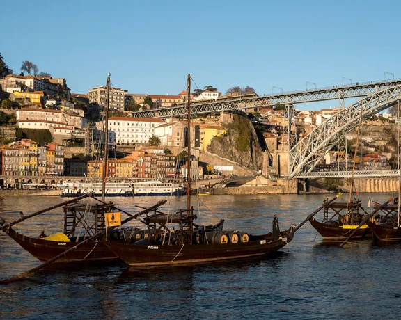 Traditional boats on a river with a cityscape and arched bridge in the background.