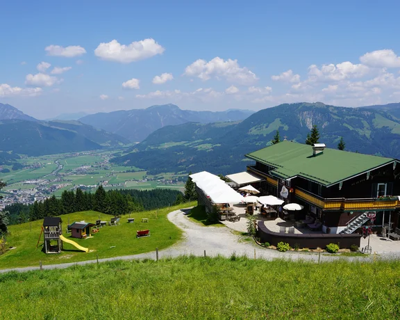Mountain lodge with green roofs overlooking a scenic valley and distant mountains under blue sky.