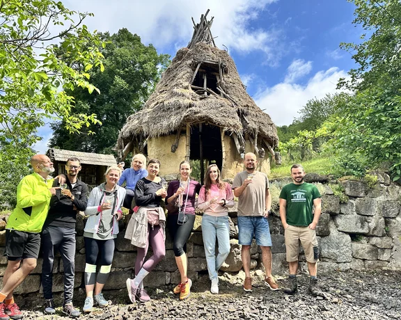 Group of people smiling in front of a small, rustic thatched-roof hut on a sunny day.