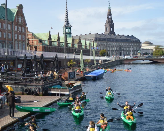 People kayaking on a canal with historic European buildings in the background.