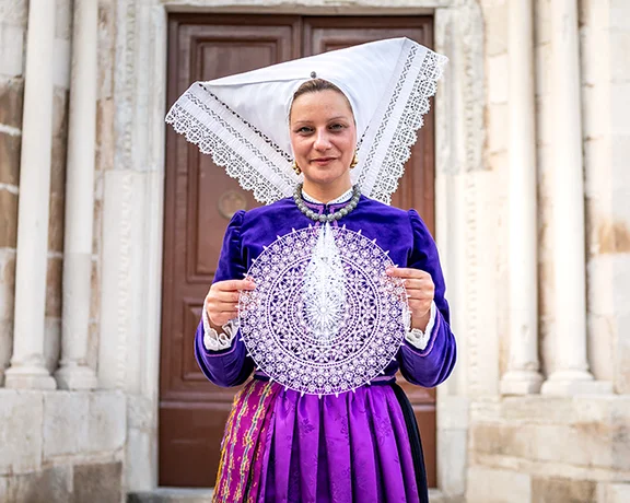 Woman in traditional purple dress and large white headdress holding lacework.