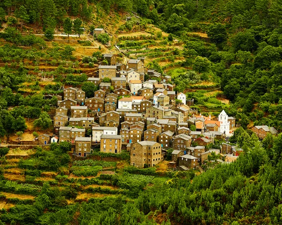 Small village with stone houses surrounded by terraced hills and green trees.