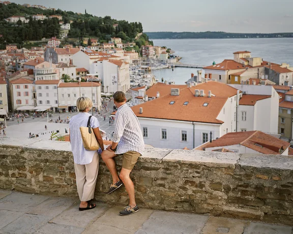 Two people lean on a stone wall overlooking a coastal town with red rooftops.