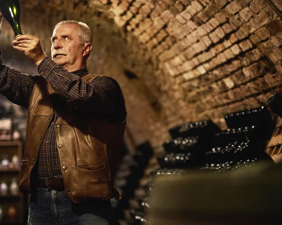 Elderly man inspecting a wine bottle in a dimly lit wine cellar.