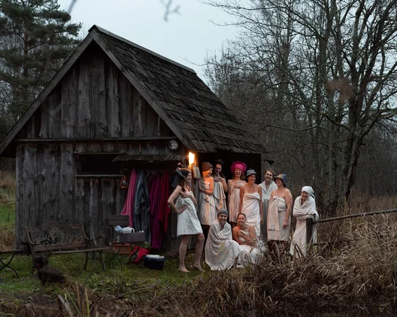 Group of people in towels gather outside a rustic wooden cabin, one holding a torch.