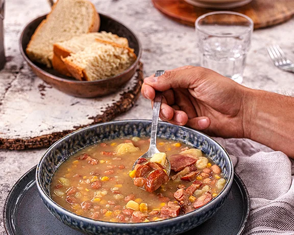 Hand holding a spoon over a bowl of hearty soup with bread on the side.