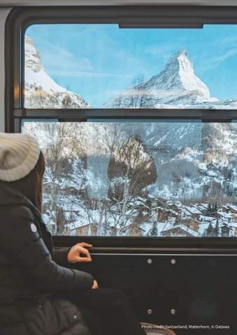 Person in winter clothing looks out train window at snowy mountains and the Matterhorn.