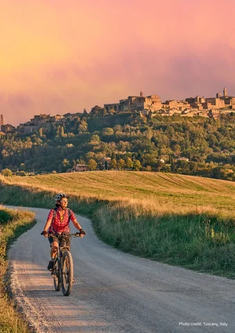 Person riding a bicycle on a rural road at sunset with a hilltop village in the background.