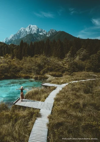 Person standing on a wooden boardwalk by a turquoise lake with mountains in the background.