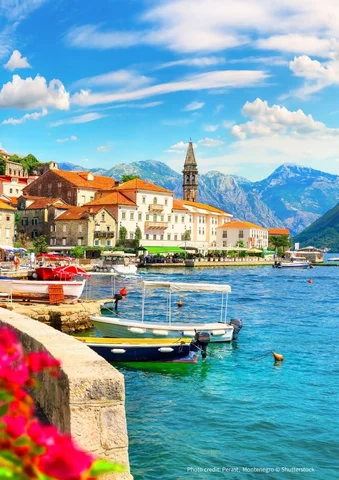 Colorful boats docked by a coastal village with red-roofed buildings and mountain backdrop.