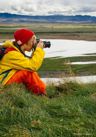 Person in a yellow jacket taking a photo by a lake with mountains in the background.