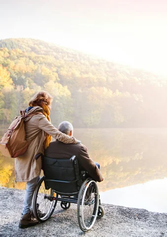 Two people by a lake, one in a wheelchair, with autumn trees in the background.