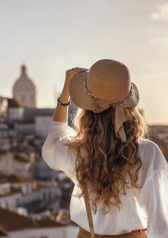 Woman with long curly hair in a hat overlooking a sunlit cityscape.