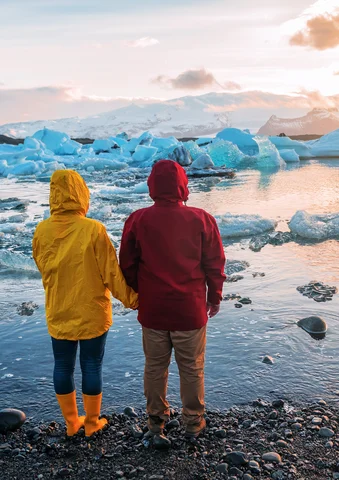 Two people in colorful jackets stand by a glacial lagoon at sunset.