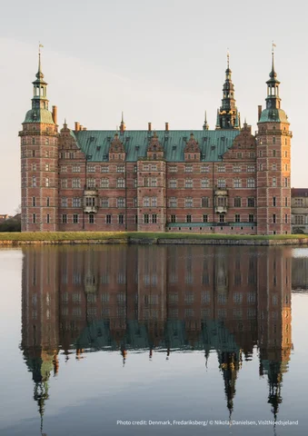 Large brick castle with green towers reflected in a calm lake at sunset.