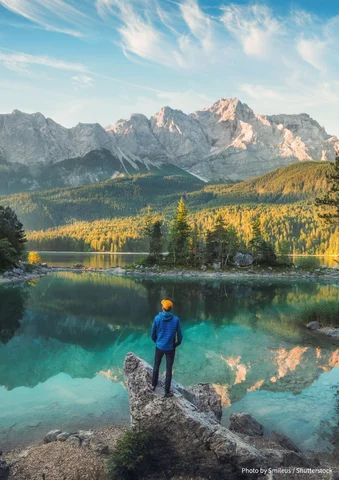 Person in blue jacket stands by a clear lake with mountains and trees in the background.