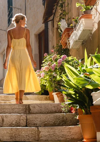 Woman in a yellow dress walks up sunlit stone steps lined with potted plants.