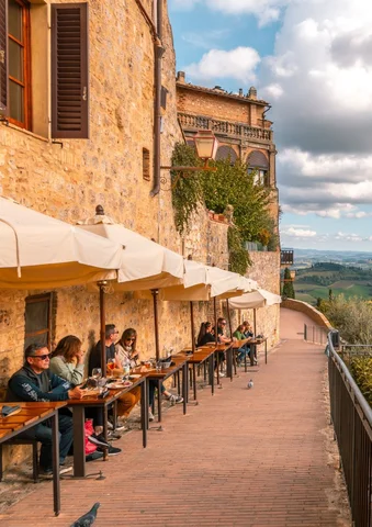 People dining outdoors along a stone building with scenic countryside views.